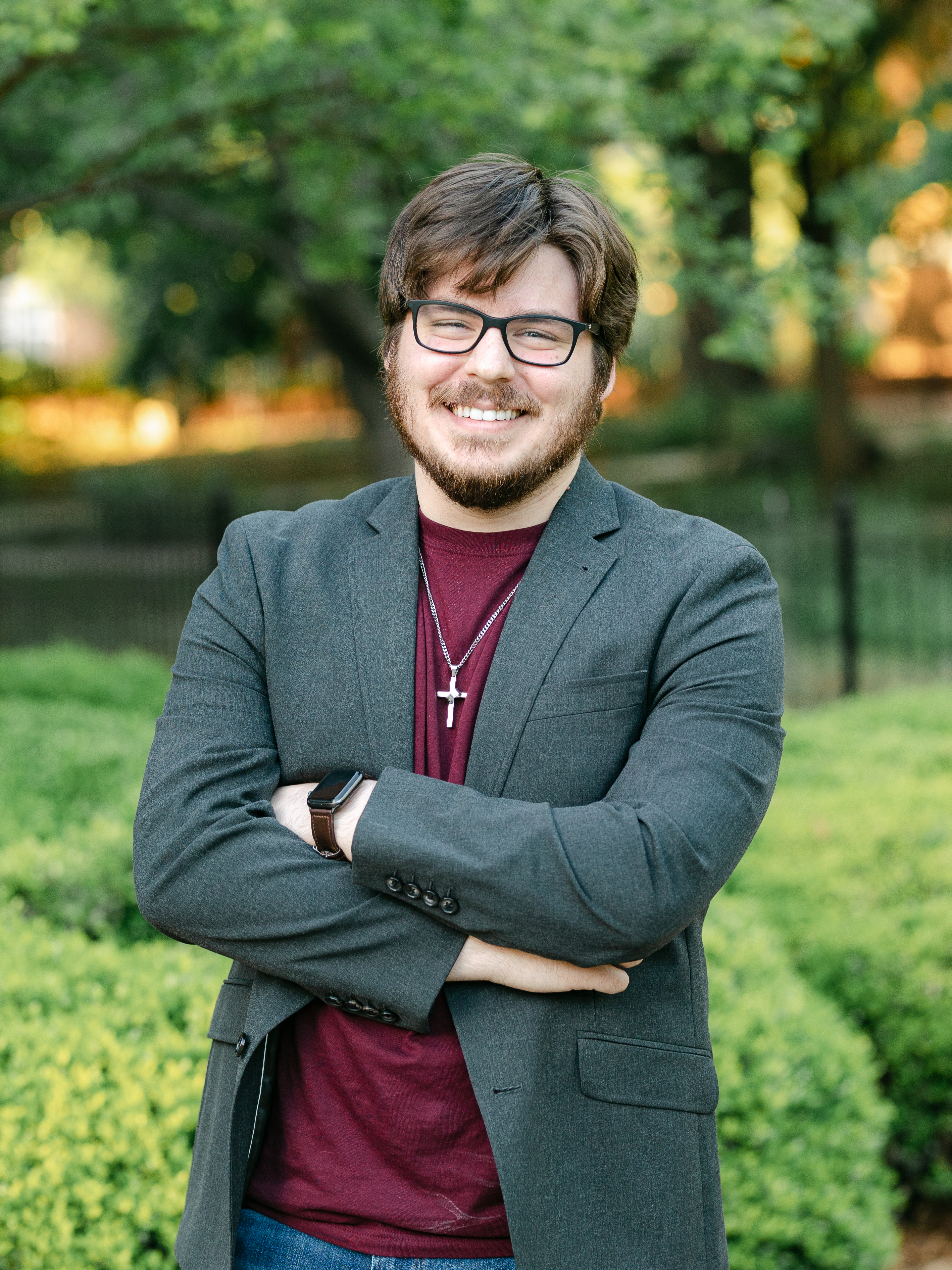 A waist-up photograph of a young white man with short brown hair, beard, and mustaache. He is wearing glasses, a maroon shirt, a dark green jacket, and is smiling.